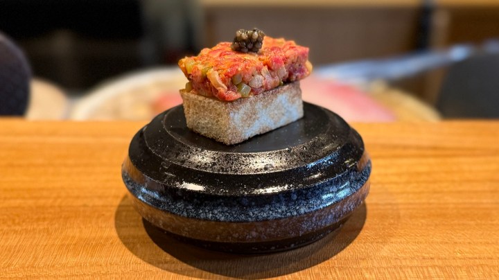 A gourmet dish featuring a small, toasted bread square topped with a vibrant tartare mixture and garnished with a dollop of caviar, presented on a circular black stone plate.