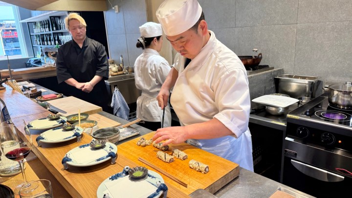 A sushi chef skillfully prepares sushi rolls at a wooden counter, while two other chefs observe in a modern kitchen setting.
