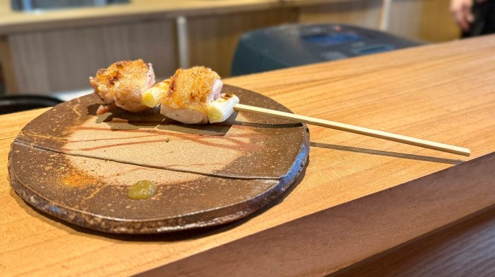 Two pieces of grilled yakitori skewers served on a round ceramic plate with chopsticks, featuring a wooden table in the background.