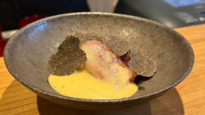 A gourmet dish served in a dark bowl featuring a piece of meat topped with black truffle slices and a creamy yellow sauce.