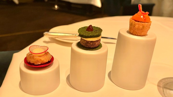 Three small gourmet desserts displayed on white pedestals, featuring a radish-topped tartlet on the left, a layered green treat in the center, and a bright orange pastry on the right.