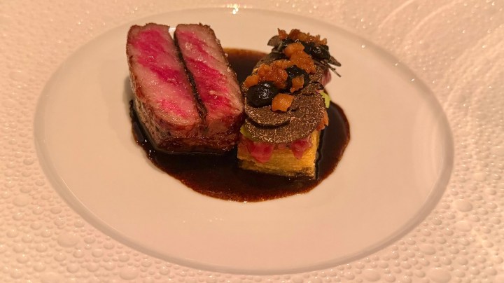 A gourmet plate featuring two slices of perfectly cooked beef, pink in the center, accompanied by a layered preparation with truffles, vegetables, and a rich sauce.