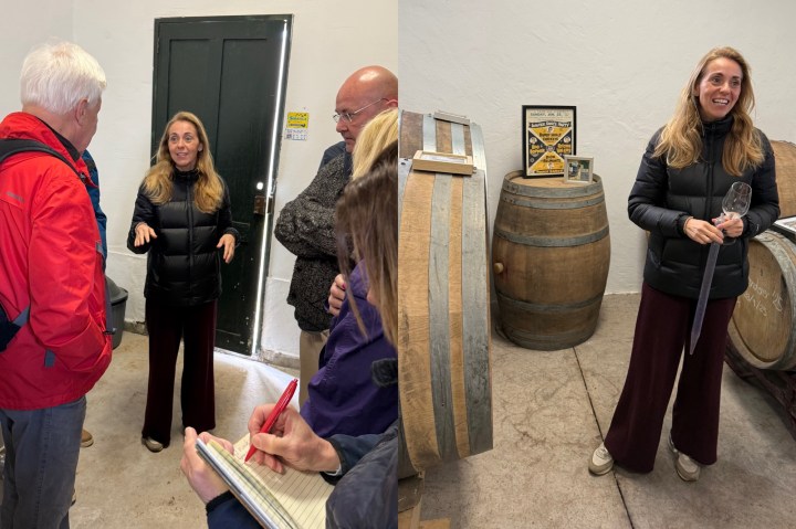 A woman in a black jacket speaks to a small group of people in a winery setting, with a wooden barrel in the background.