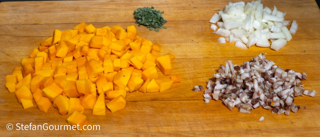 Chopped butternut squash, diced onion, and minced bacon on a wooden cutting board, with a sprinkle of fresh herbs.