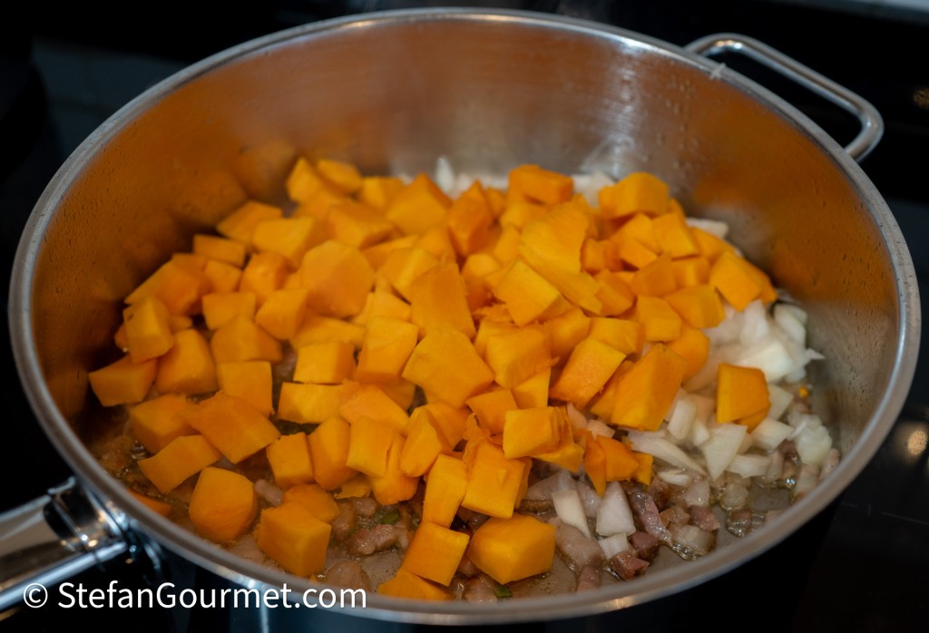 A close-up of a pot containing diced butternut squash and chopped onions being sautéed, with some bits of meat visible at the bottom.