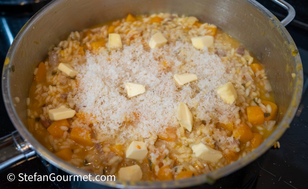 A pot of creamy risotto featuring chunks of butternut squash, topped with grated cheese and small cubes of butter, cooking on a stovetop.