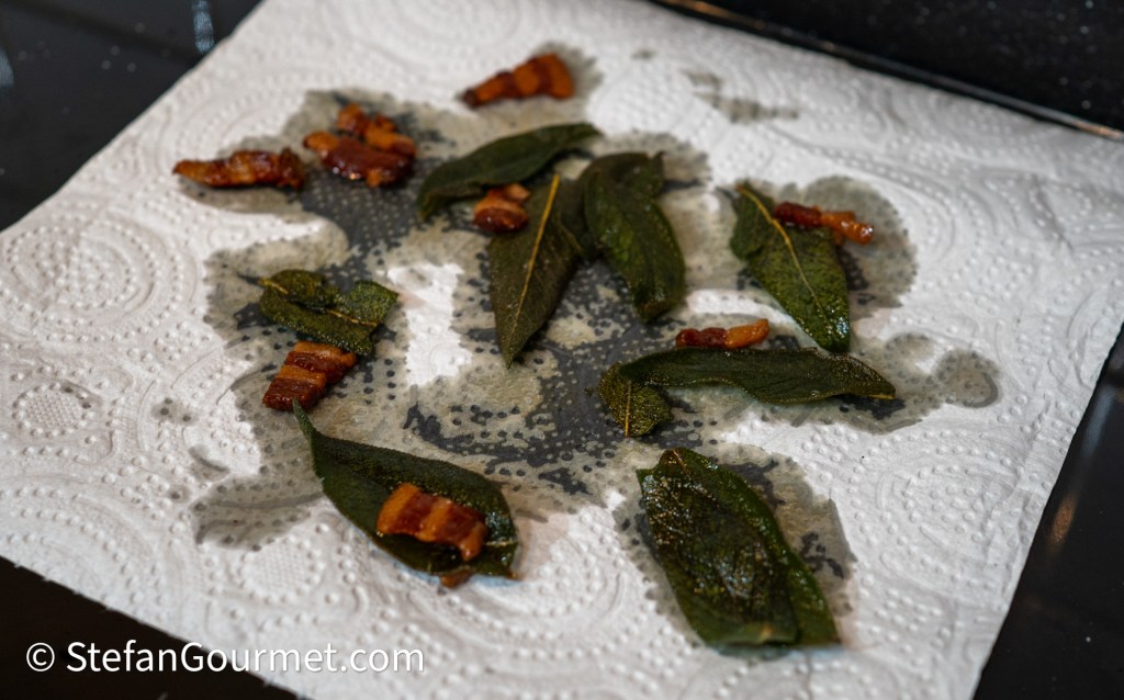 Crispy fried sage leaves and small pieces of bacon resting on a paper towel.