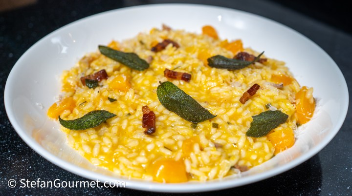 A creamy risotto dish topped with crispy sage leaves, chunks of butternut squash, and bits of bacon, served in a white bowl.