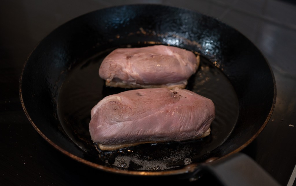 Two pieces of meat cooking in a frying pan with some oil.