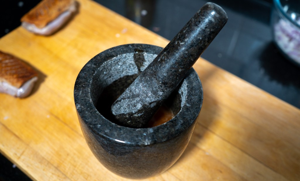 A granite mortar and pestle on a wooden cutting board, with pieces of meat in the background.