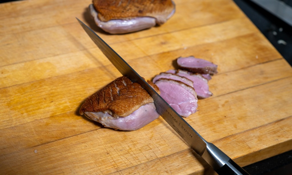 A chef slicing cooked duck breast on a wooden cutting board, with a sharp knife.
