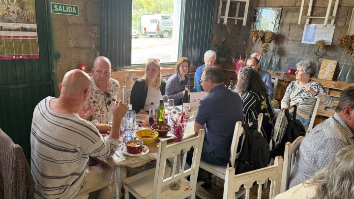 A group of people gathered around a table in a rustic setting, enjoying food and drinks. The table is adorned with various dishes and bottles, while the background features green windows and walls decorated with dried flowers.