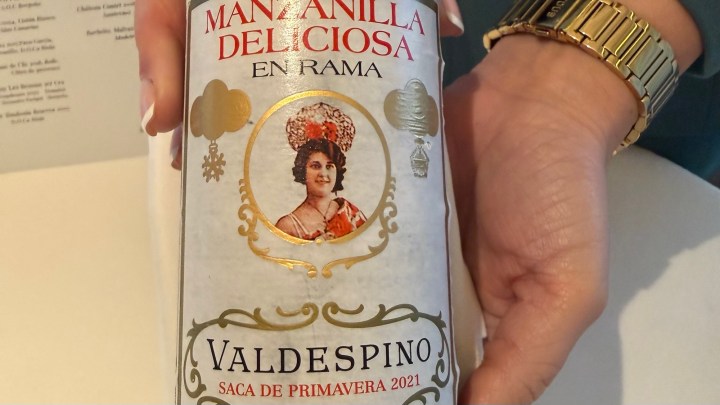 A close-up of a hand holding a bottle of Valdespino Manzanilla Deliciosa en Rama, featuring a decorative label with an illustration of a woman and gold accents, indicating it is from the Spring 2021 selection.