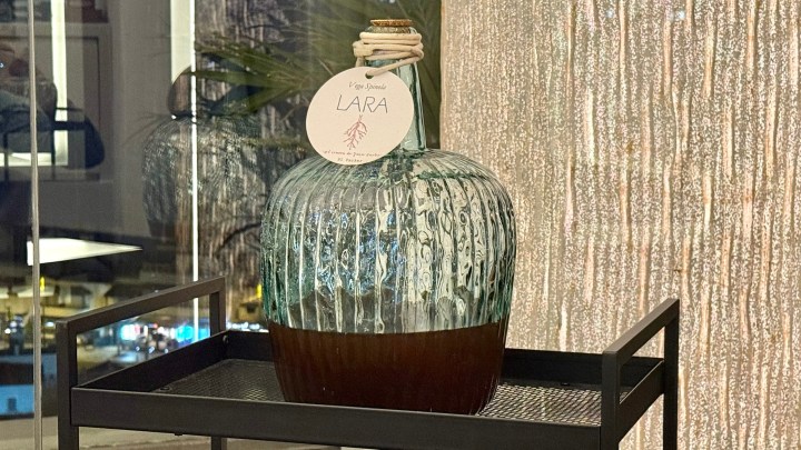 A decorative large glass bottle with a textured design, filled with a dark liquid and topped with a label reading 'LARA'. The bottle is placed on a black metal tray with a mesh bottom.