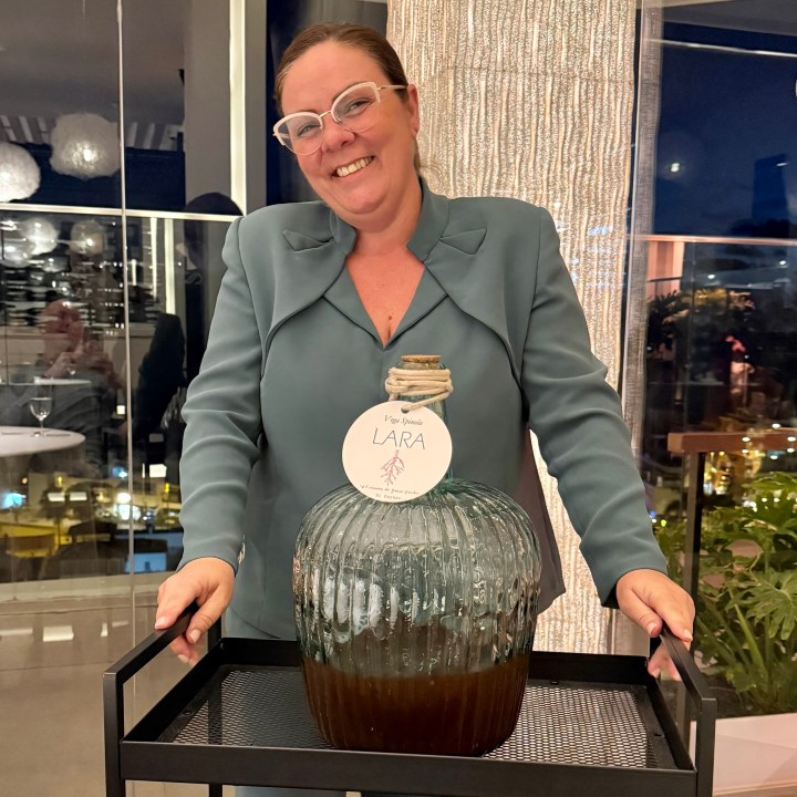 A woman smiling and standing behind a glass container filled with a dark liquid, labeled 'LARA', on a tray in a sophisticated setting.