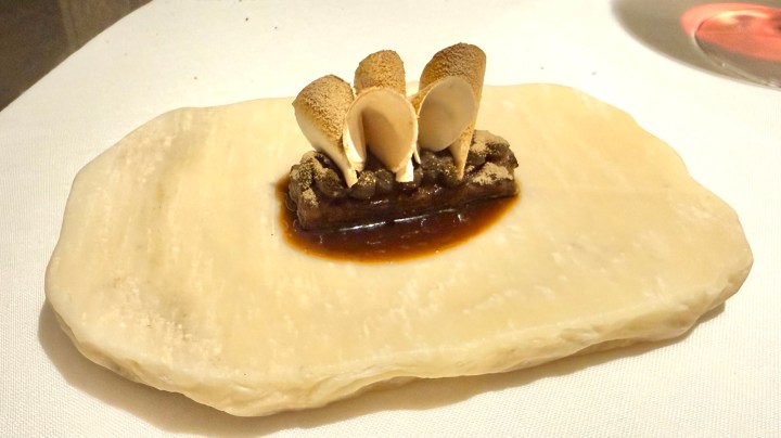 An artistic presentation of a gourmet dish featuring a small rectangular piece topped with four cone-shaped elements, garnished with a glossy sauce, served on a large, smooth, and light-colored plate.