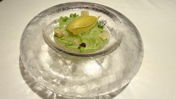 An elegant dish presented in a textured glass bowl featuring a yellow sorbet atop a bed of green shaved ice, surrounded by small pieces of bread and drizzled with green sauce and garnished with herbs.