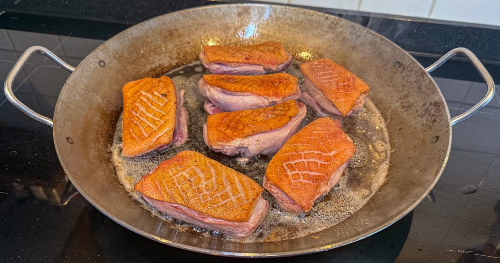 Eight pieces of seared duck breast cooking in a large frying pan with a golden-brown crust and sizzling in oil.