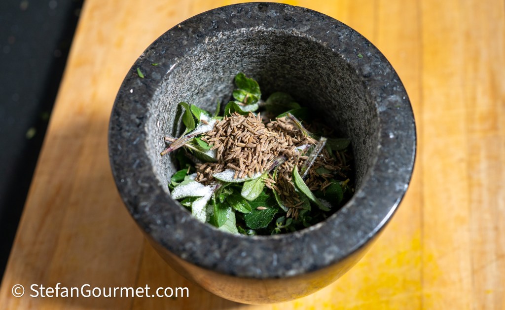 A close-up of a mortar and pestle containing fresh herbs and spices, including green leaves and cumin seeds, set on a wooden surface.