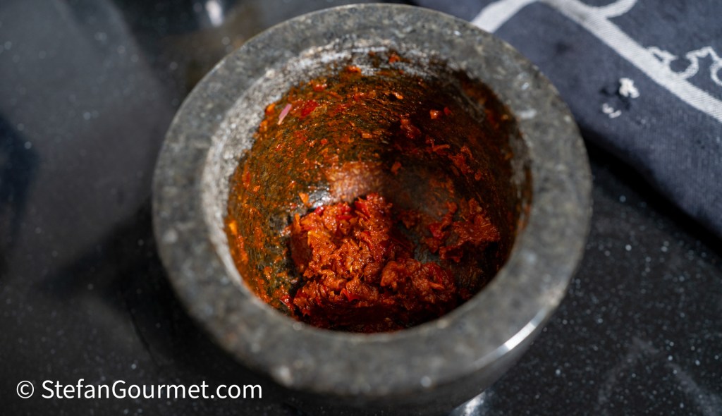 A stone mortar filled with a red chili paste, with a textured surface and a dark countertop in the background.