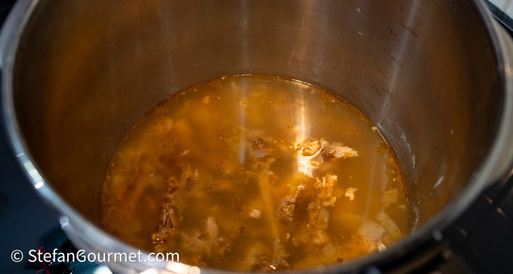 Close-up of a stainless steel pot filled with a brown liquid and small pieces of meat, indicating the preparation of a soup or broth.