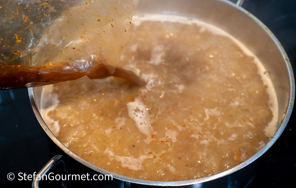 A person pouring a thick, brown sauce from a plastic bag into a simmering pot of soup or stew.