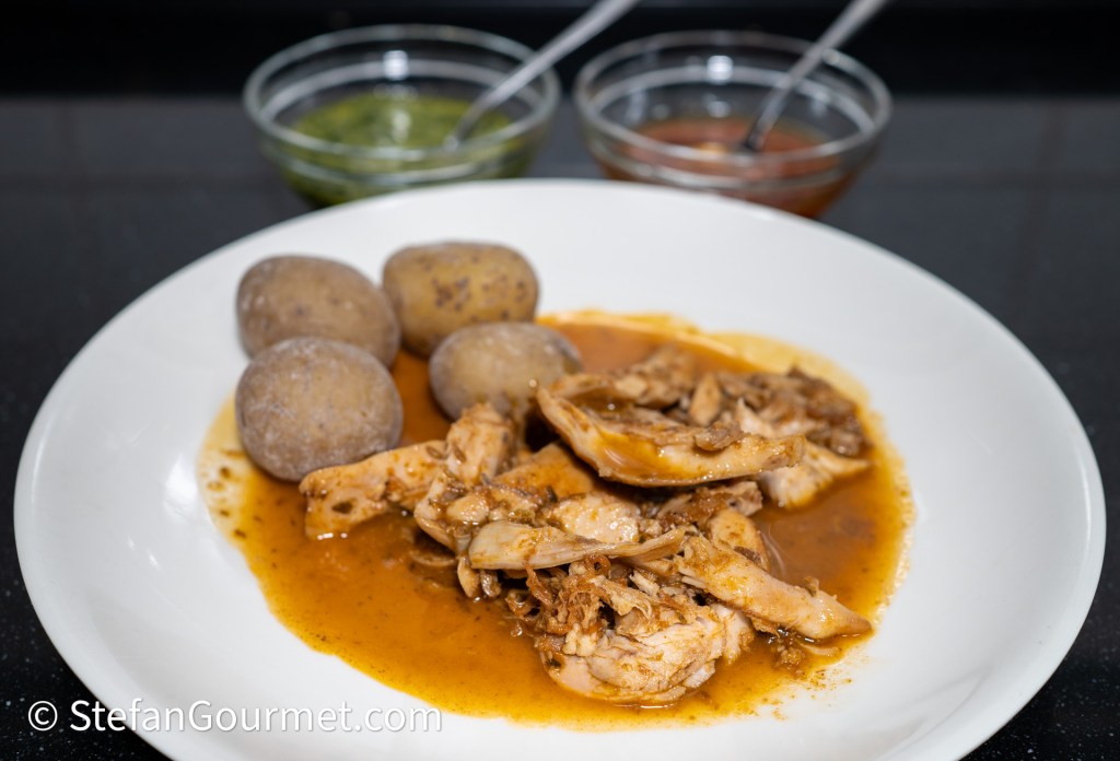 A plate of tender chicken pieces in a savory sauce, accompanied by small boiled potatoes and two bowls of green and red sauces.