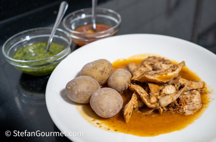 A plate of shredded chicken in sauce served with small boiled potatoes, accompanied by two bowls of green and red sauces.