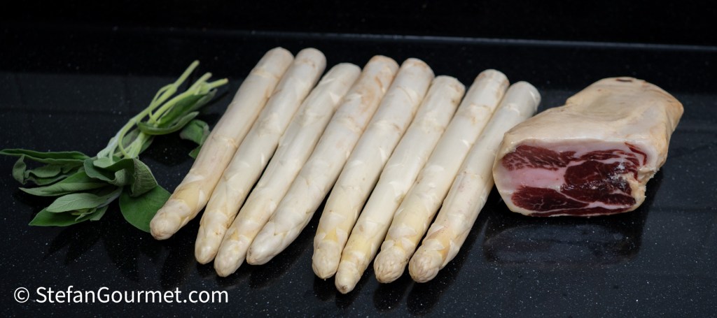 A bunch of white asparagus spears next to a piece of cured meat and fresh sage leaves on a black countertop.