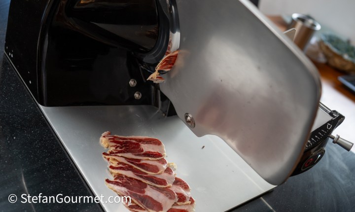 A meat slicer with freshly sliced cured meat on a stainless steel surface.