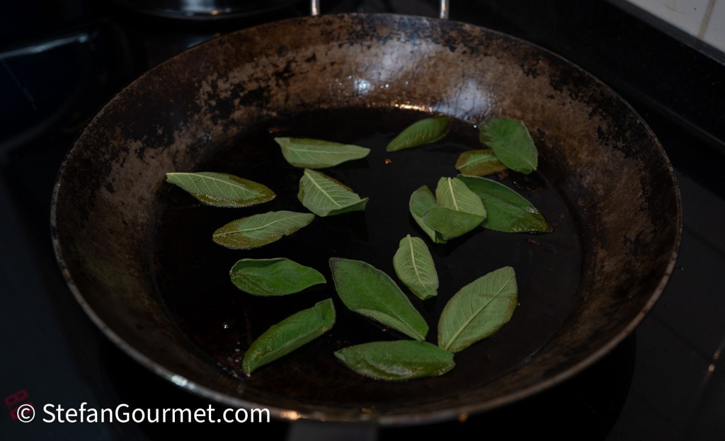 A frying pan with several fresh bay leaves floating in oil, placed on a stovetop.