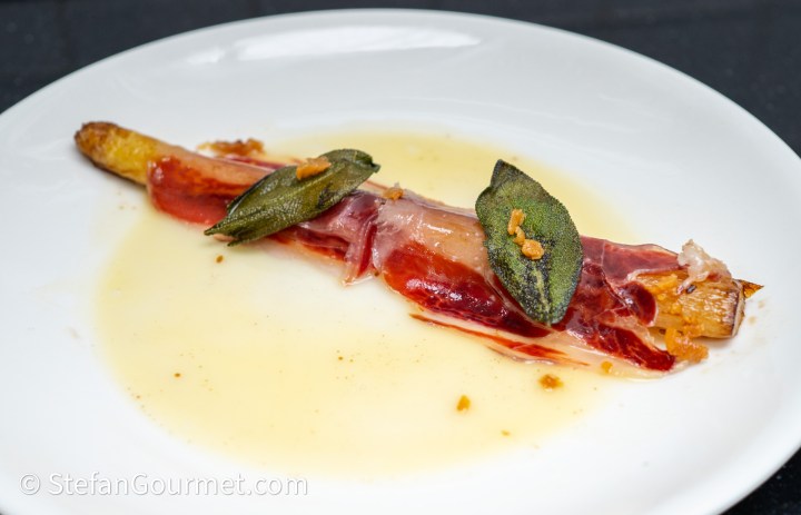 A plated dish featuring a piece of asparagus wrapped in prosciutto, garnished with sage leaves and a drizzle of sauce.