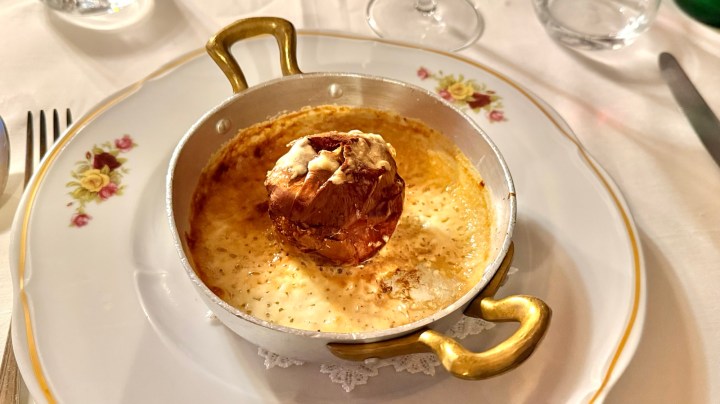 A small silver serving dish with a golden brown baked dish topped with cheese and an onion in the center, placed on a decorative plate.