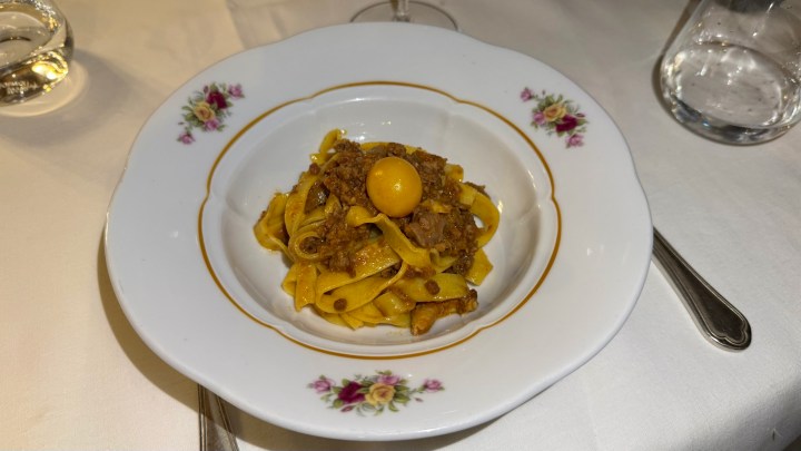 A plate of tagliatelle pasta topped with a cooked meat sauce and a raw egg yolk, garnished with herbs, served on a vintage floral plate.