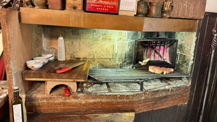 A rustic indoor grill with a stainless steel surface, featuring a piece of grilled meat, cooking on a charcoal fire, surrounded by cooking utensils and condiments.