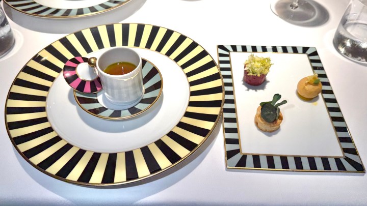 An elegantly presented dish featuring a large round plate with a striped design, holding a small cup with liquid, and a smaller rectangular plate displaying three gourmet appetizers.