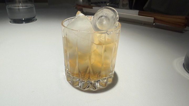 A glass of whiskey on the rocks with two ice cubes, placed on a textured white surface.