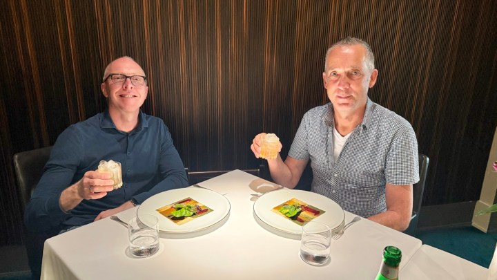 Two men sitting at a table in a restaurant, each holding a drink, with dishes of colorful food in front of them.