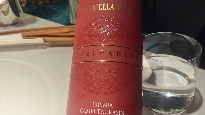 A close-up of a red wine bottle labeled 'Trespadini Irpinia Campi Taurasini' with decorative embossing, placed next to a glass of water.