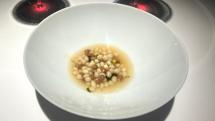 A white bowl containing small round pasta-like beads in a light broth, accompanied by garnish, with two glasses of red wine in the background.