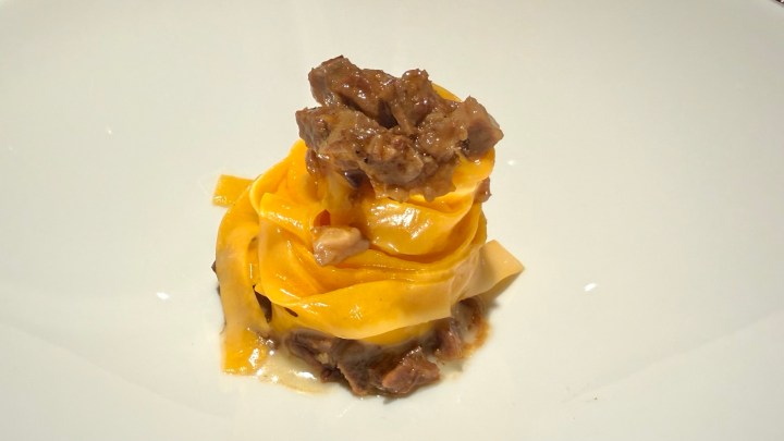 A serving of yellow pasta twirled into a nest, topped with pieces of meat and a rich sauce, presented on a white plate.