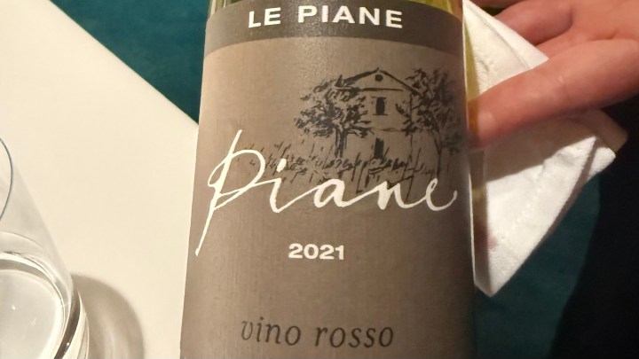 A close-up of a wine bottle labeled 'Le Piane 2021 Vino Rosso' with a rustic illustration of a house and tree on the label.