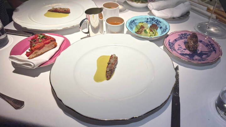 A gourmet dining table featuring a white plate with a single piece of food drizzled with olive oil, accompanied by a pink plate with a slice of toast topped with greens, a blue plate with salad, and a pink plate with a decorative food item. Two cups are visible, one with a dark beverage, and another made of silver.