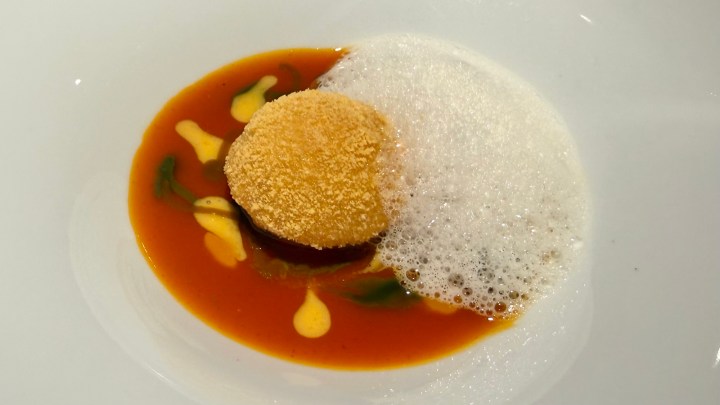 A gourmet dish featuring a round, textured ball sitting in a vibrant orange sauce, surrounded by frothy bubbles and colorful garnishes.