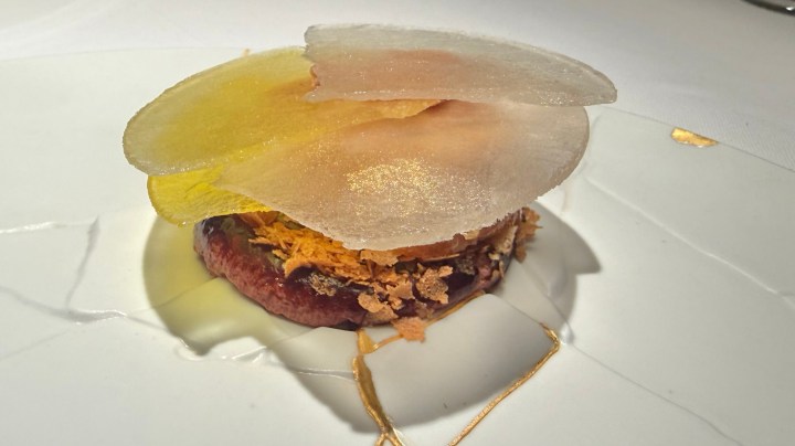 An artistic dish featuring layered elements, including translucent yellow and white components, atop a base of crumbled textures, served on a white plate.