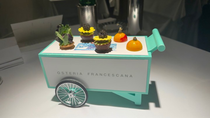 A decorative dessert cart with a turquoise finish, labeled 'OSTERIA FRANCESCA,' featuring various intricate desserts including miniature cakes adorned with flowers and fruits.