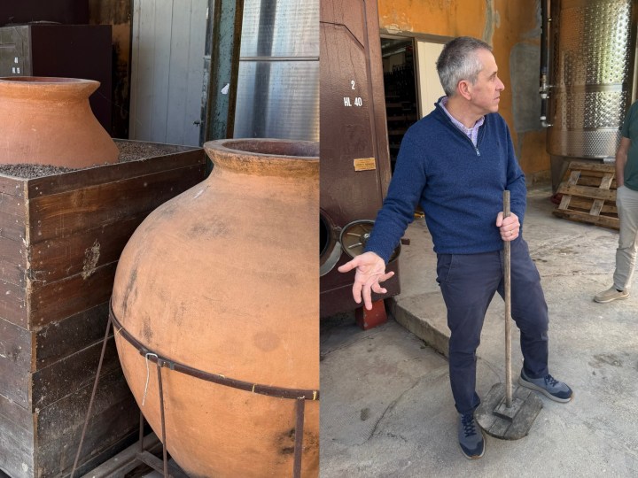 A large terracotta pot next to a wooden box filled with dark material, alongside a man in a blue sweater gesturing while holding a tool, in a winery setting.