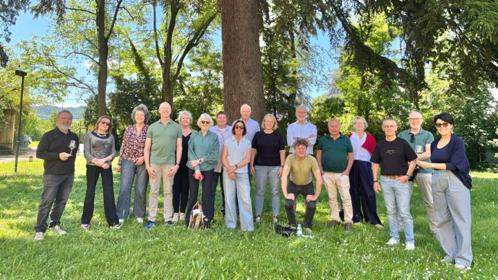 Group photo of 20 adults standing together in a grassy park area, surrounded by trees.