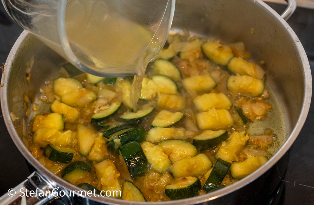 A pot containing diced zucchini and chunks of squash cooking with broth being poured in.