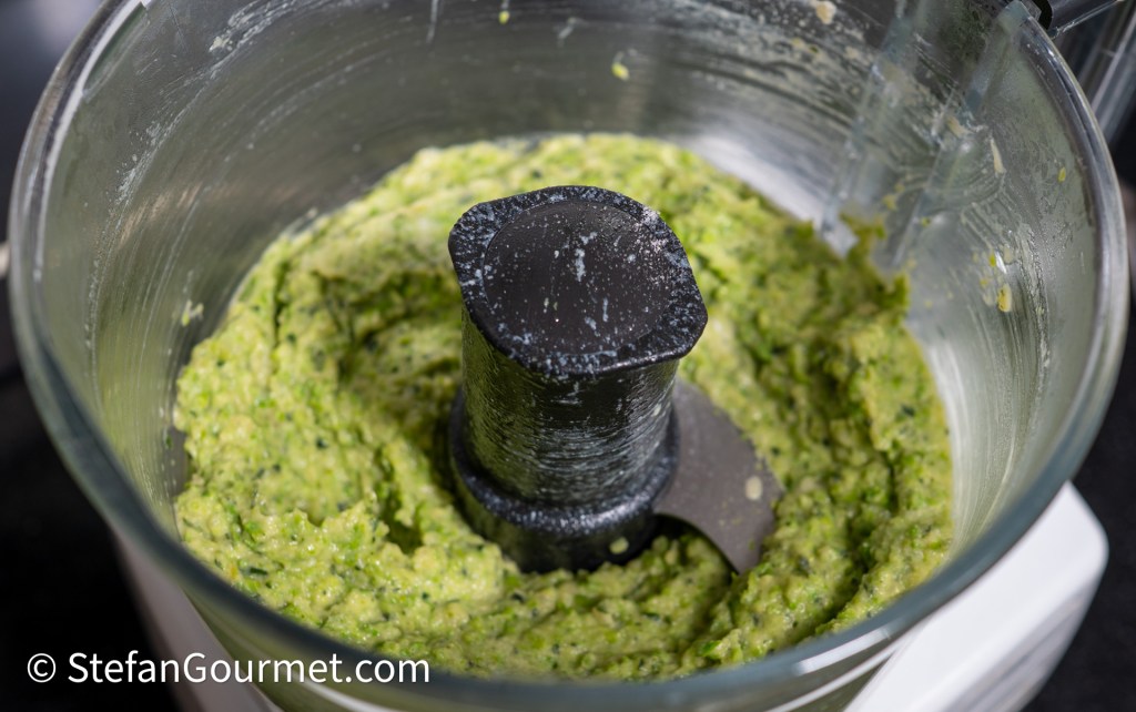 A top view of a food processor containing green blended mixture, showing the blade in the center.